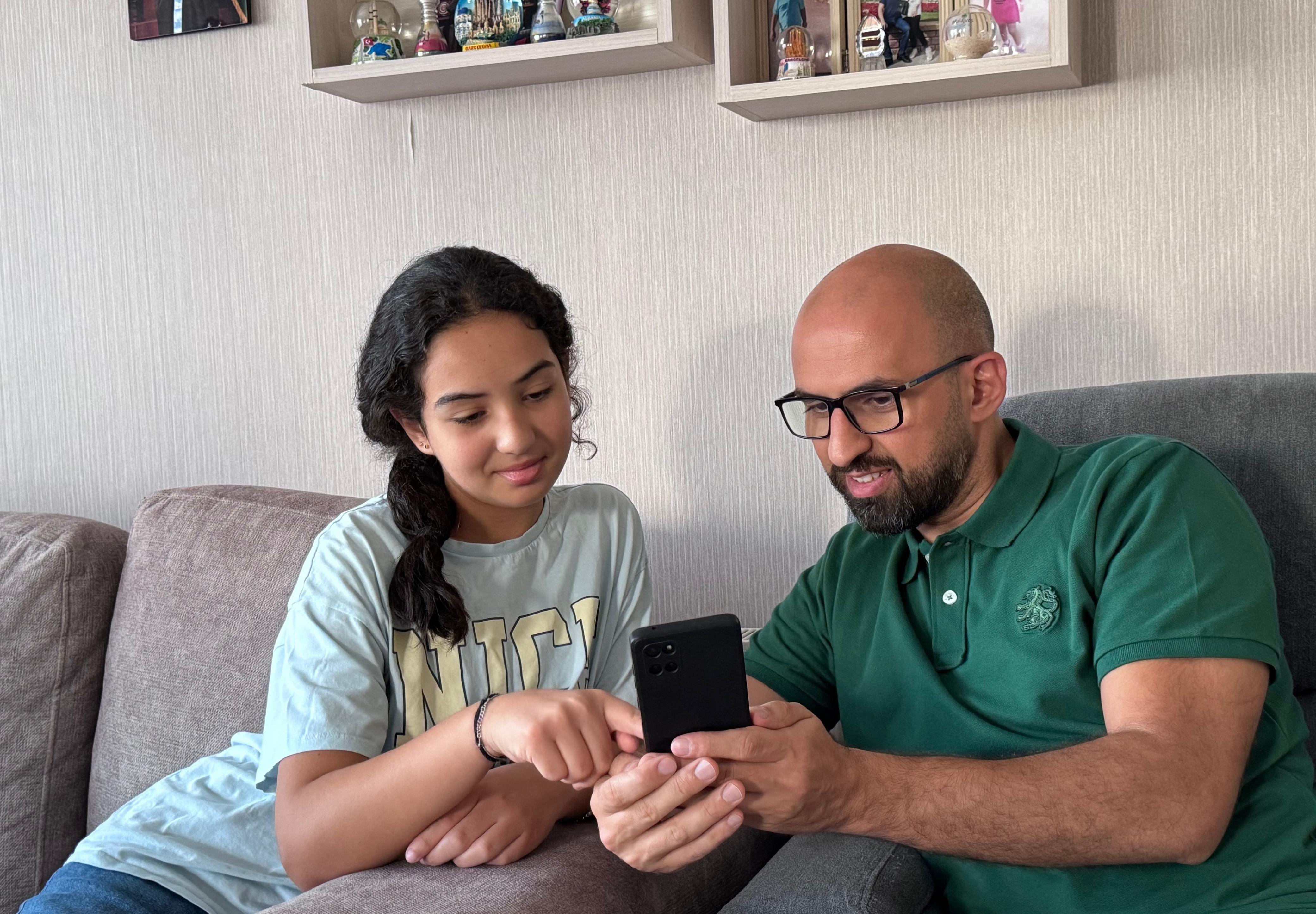 عمر يلبس قميصاً أخضر ويمسك بهاتفه ويجلس مع ابنته ليان يريها شيئاً على الهاتف