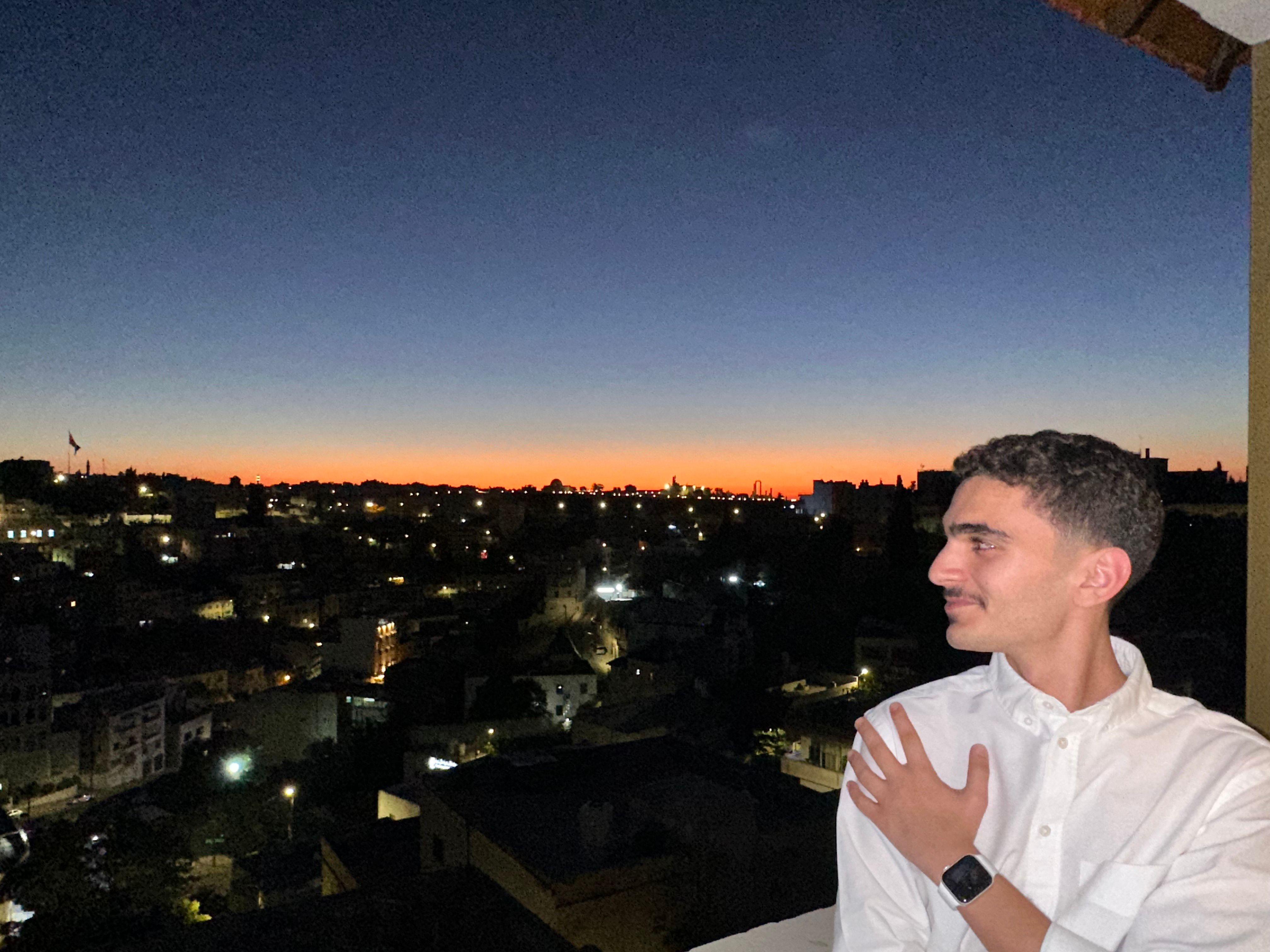 صورة لمحمد أديب (21 سنة) طالب إدارة، يرتدي قميص أبيض وتظهر خلفه مدينة عمّان
