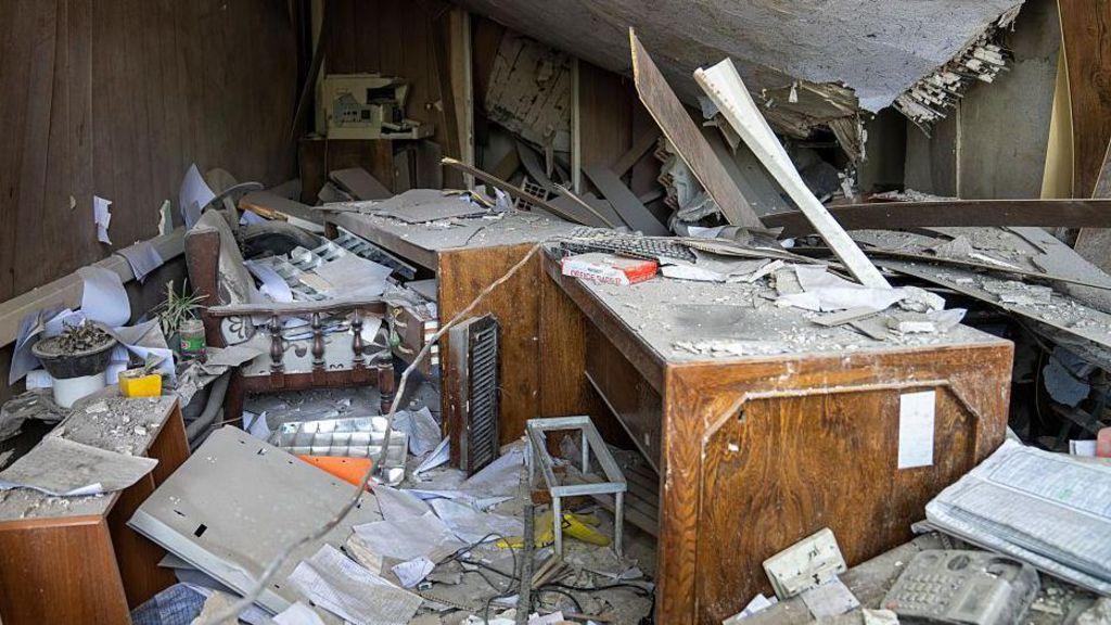 معدات مكتبية متضررة وسط الدمار في سجن إيفين بعد قصفه بغارة جوية إسرائيلية. انهار السقف وتناثرت أكوام من الأوراق. يوجد مكتب خشبي في وسط الغرفة.