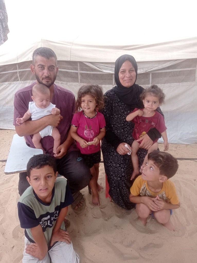 منذر يحمل بيده طفلاً رضيعاً وبجانبه زوجته التي تحمل طفلتها الصغيرة. وفي الصورة أيضاً طفلته الكبرى وطفلاه الأكبر سناً. يجلسون جميعاً في خيمة.